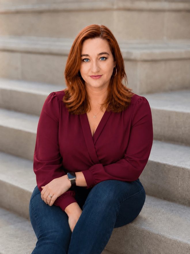 Coach Cinnamon B, survivor, sexologist, and somatic intimacy coach, sitting on stone steps in a burgundy blouse